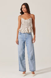 A woman is wearing a cream-colored sleeveless top with dark brown polka dots, featuring thin straps and a ruffled hem. She has paired it with light blue, high-waisted, wide-leg jeans. She is also wearing white pointed-toe shoes.